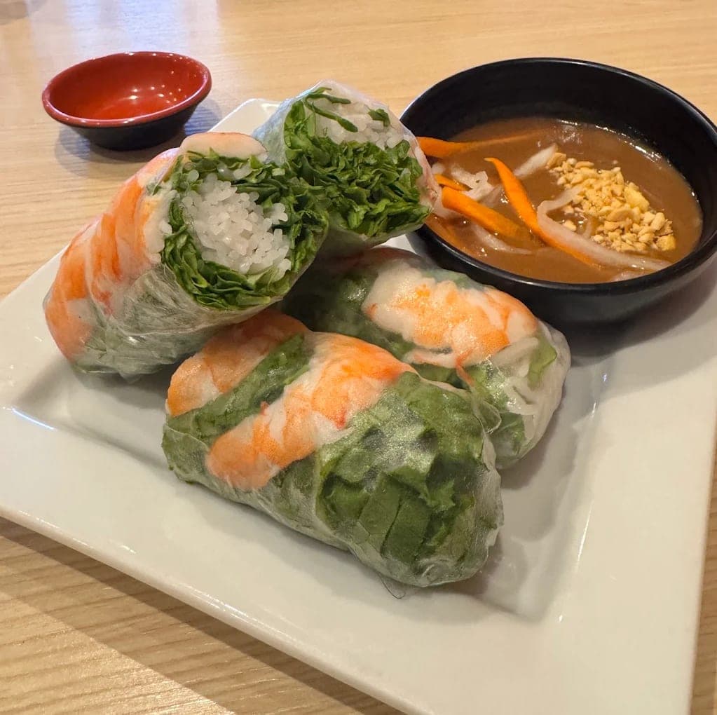 Shrimp summer rolls on a white plate, one cut open to show rice noodles and herbs, beside a bowl of peanut sauce topped with crushed peanuts