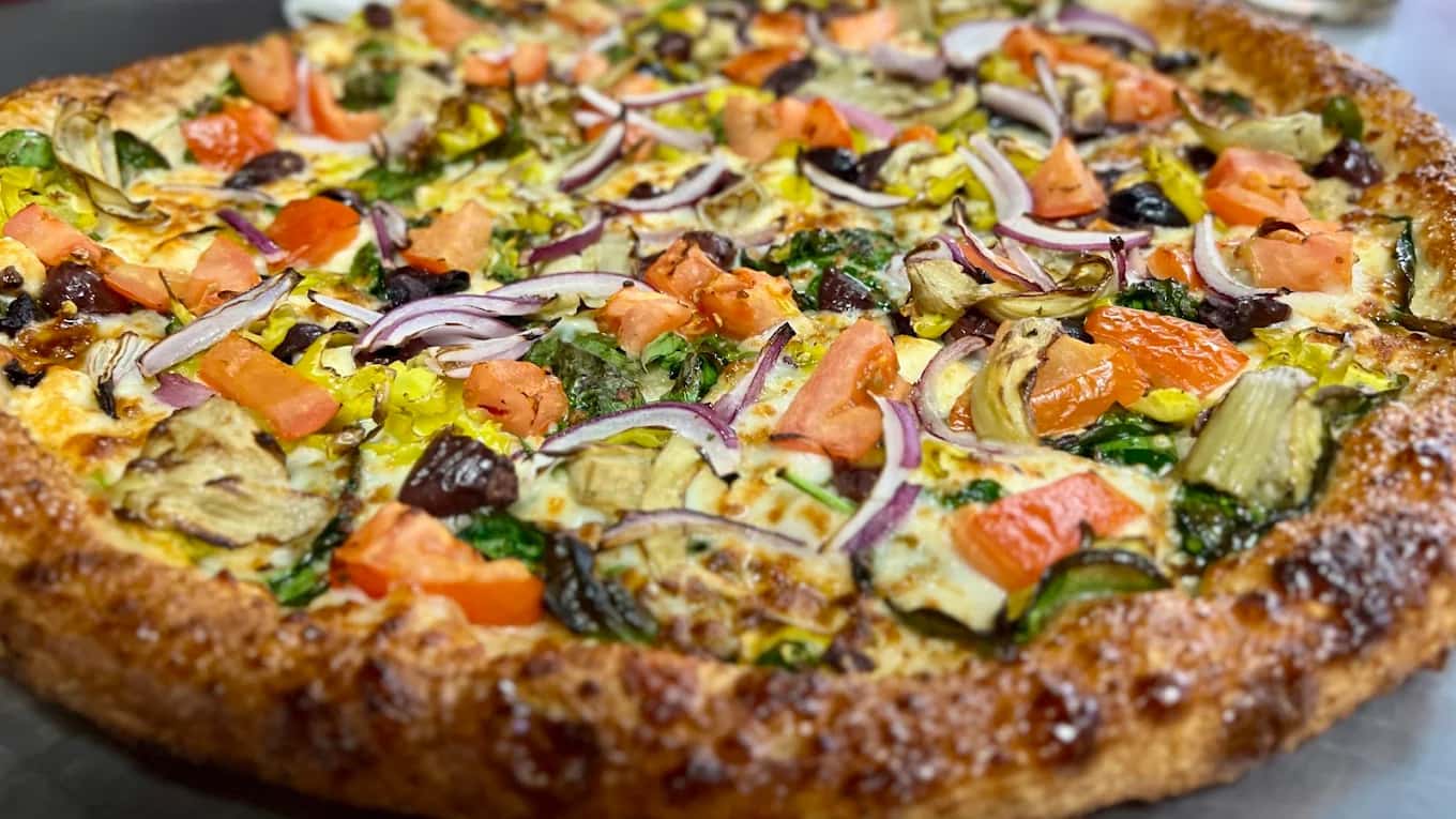Close-up of a 19th Street Pizzeria pizza topped with tomato, red onion, spinach, artichoke and olives
