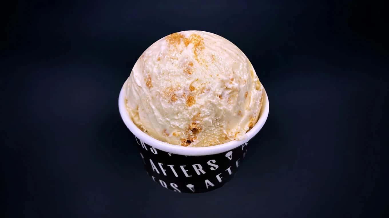 A scoop of pale, caramel-swirled ice cream heaped over the rim of a black Afters cup against a dark background
