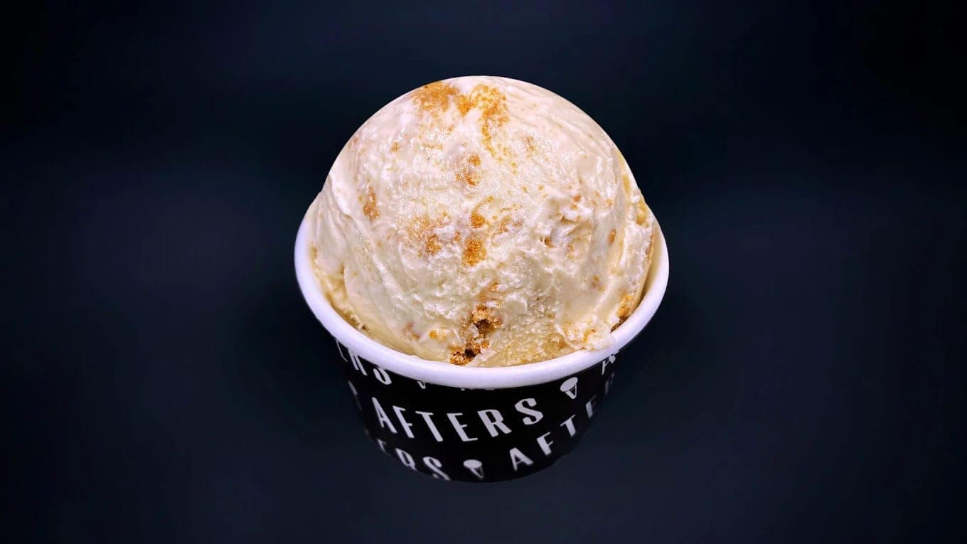 A scoop of pale, caramel-swirled ice cream heaped over the rim of a black Afters cup against a dark background