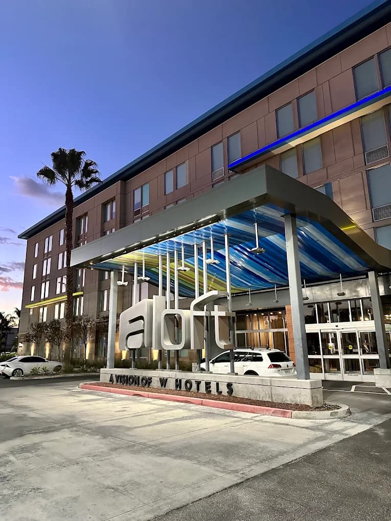 The Aloft hotel entrance at dusk, its lit lettering beneath a canopy patterned in blue and yellow, a palm tree alongside