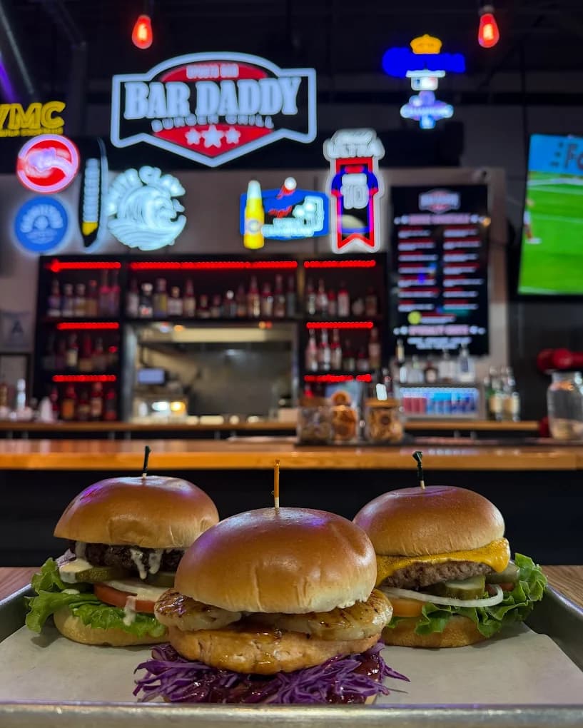 Three burgers on a tray in front of the bar and neon signs at Bar Daddy Burger & Grill