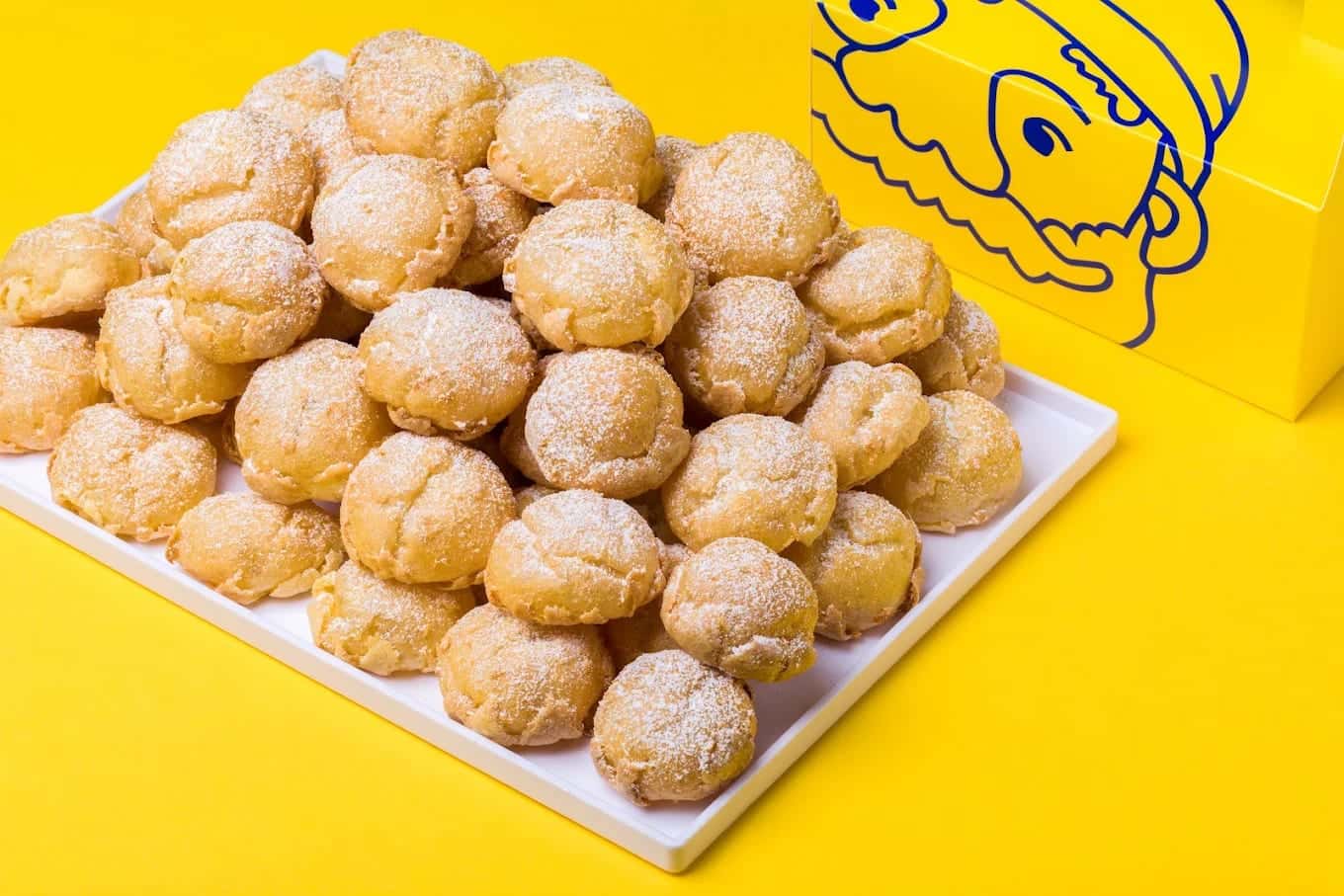 A white tray heaped with sugar-dusted cream puffs on a yellow background, beside a box carrying the Beard Papa face