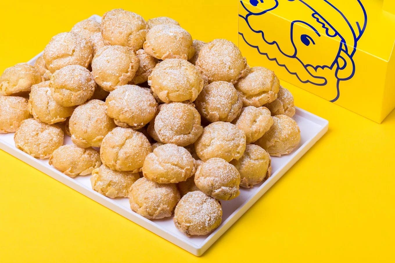 A white tray heaped with sugar-dusted cream puffs on a yellow background, beside a box carrying the Beard Papa face