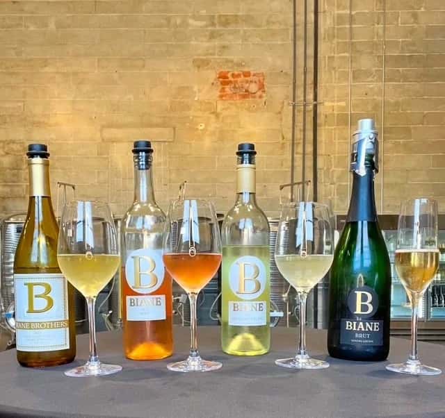 Four bottles of Biane Winery wine with filled glasses lined up on a table in front of a brick wall