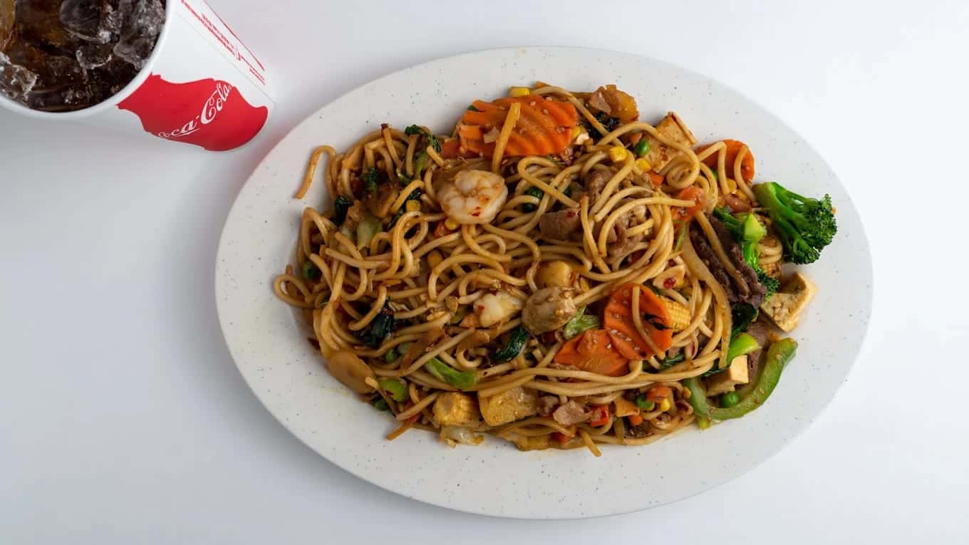 A plate of stir-fried noodles with shrimp, carrot, broccoli and tofu, a fountain drink alongside
