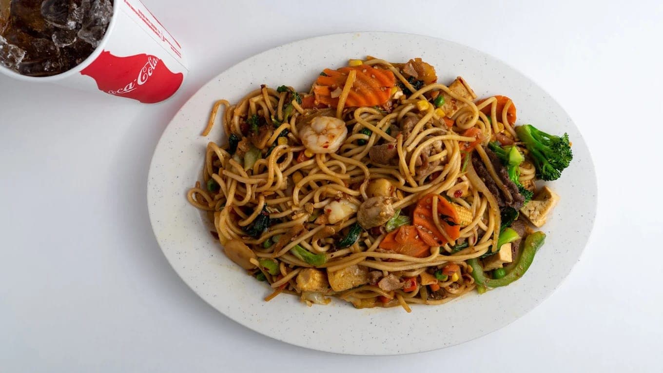 A plate of stir-fried noodles with shrimp, carrot, broccoli and tofu, a fountain drink alongside