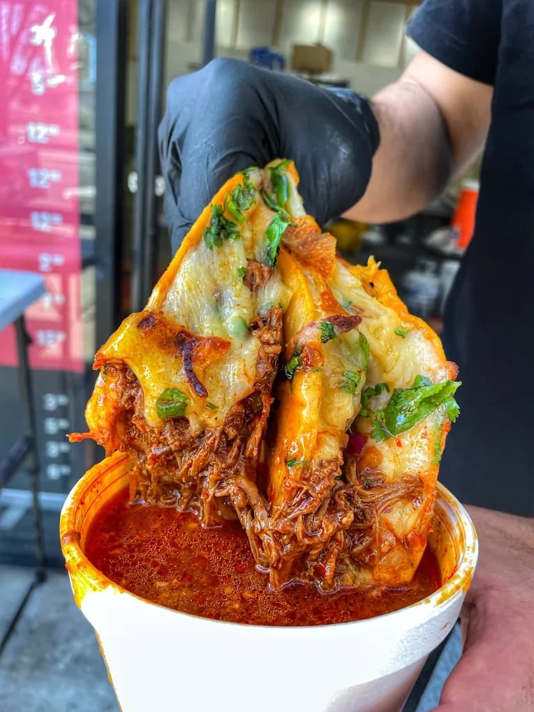 Gloved hand holding cheesy shredded beef quesatacos over a cup of red consomé at Birria Los Socios