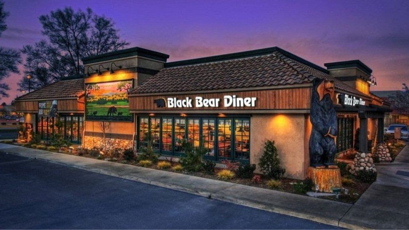 The Black Bear Diner at dusk, its lit sign on a log-cabin front with a carved wooden bear standing by the door