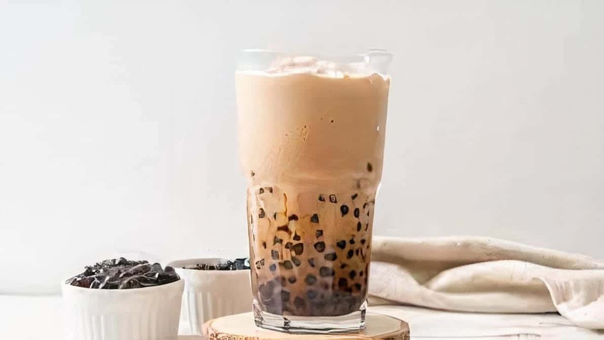 A tall glass of milk tea with tapioca pearls settled at the base, small dishes of pearls beside it on a wooden coaster