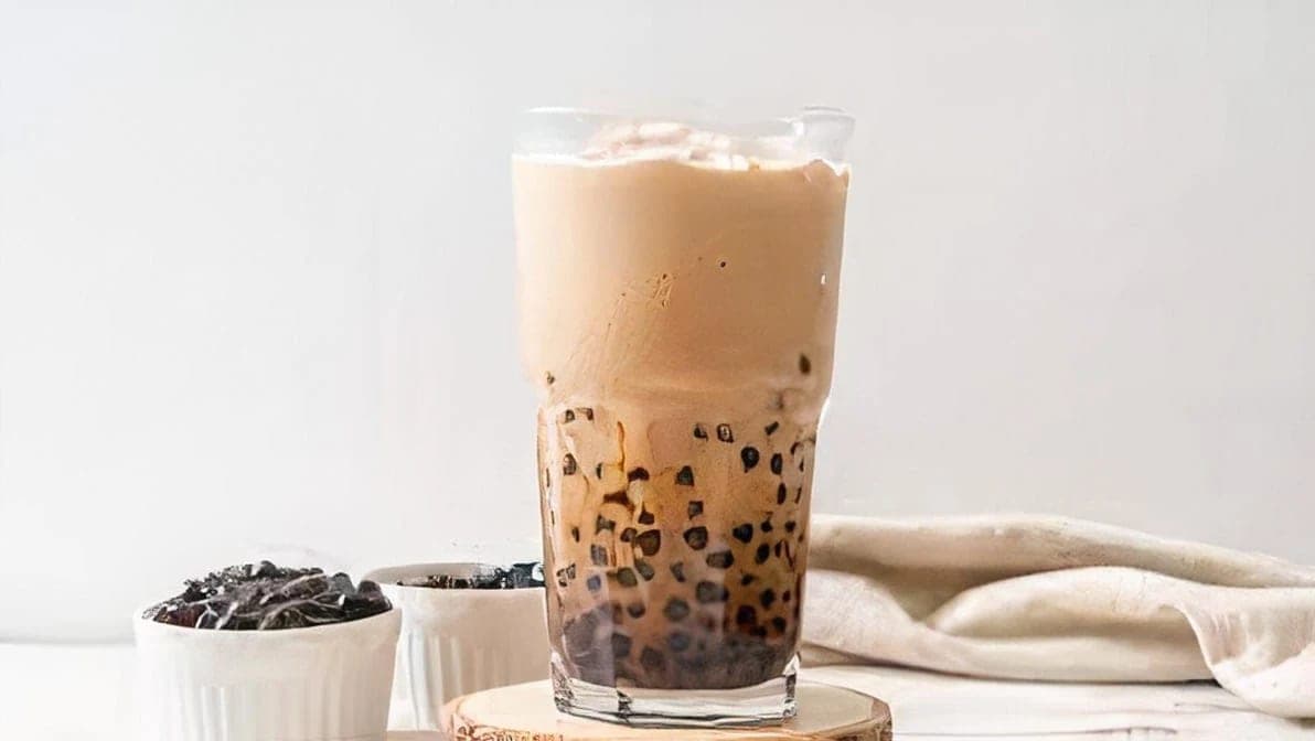 A tall glass of milk tea with tapioca pearls settled at the base, small dishes of pearls beside it on a wooden coaster