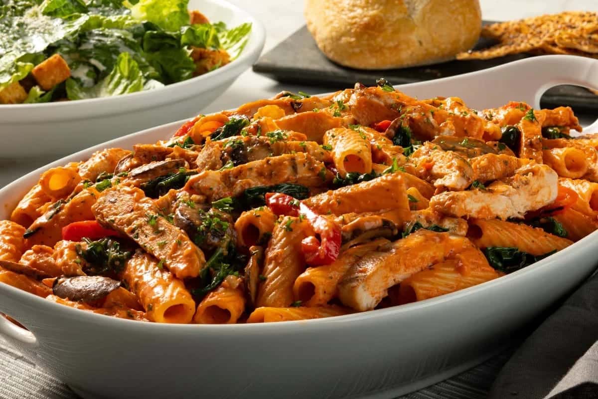 An oval dish of rigatoni in tomato cream with chicken, spinach and mushrooms, a Caesar salad and bread behind it