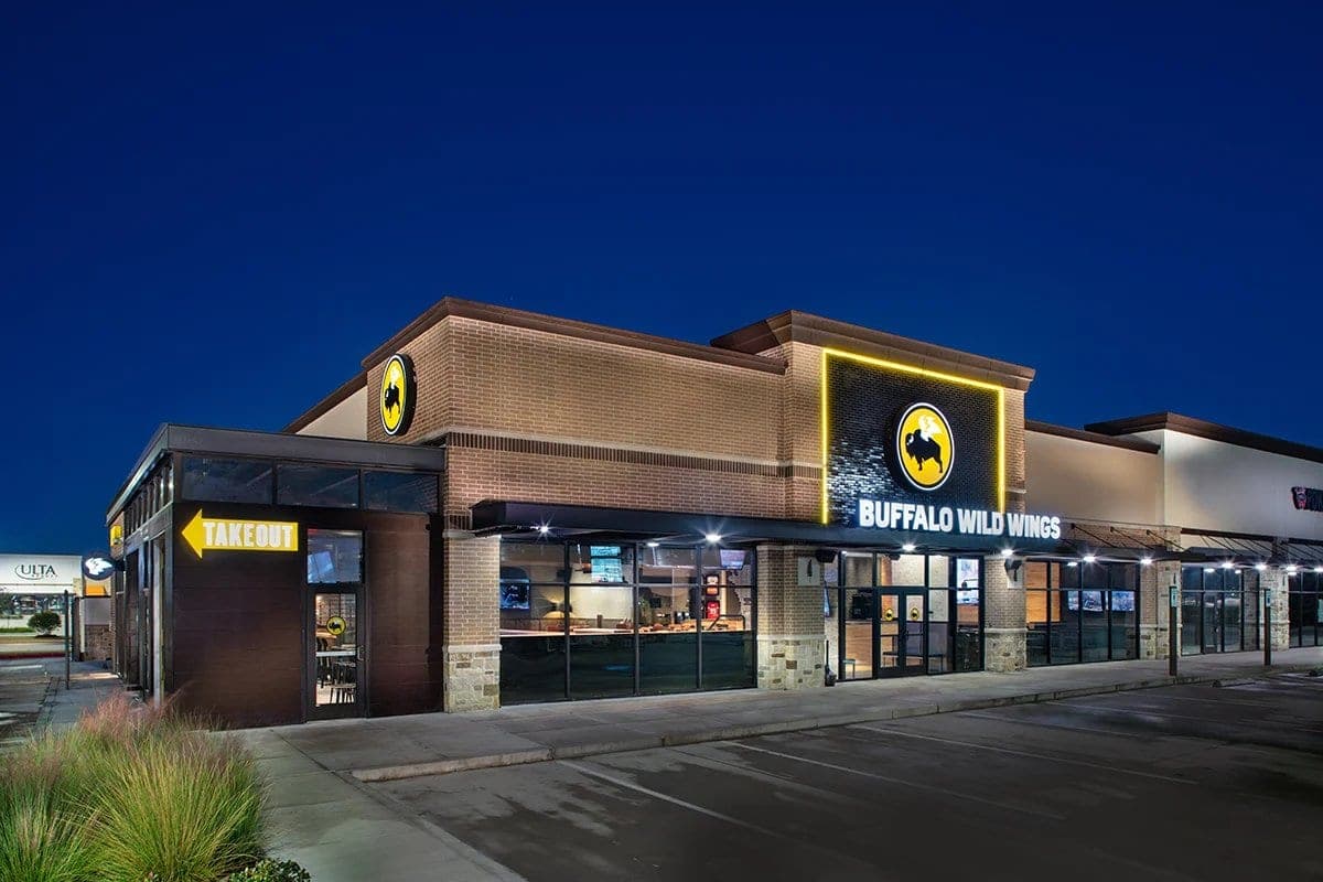 The Buffalo Wild Wings exterior at night, its lit buffalo logo above a brick facade with a takeout entrance at one end