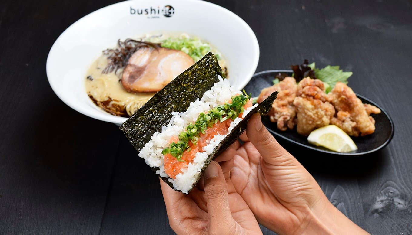 Hands holding a salmon rice hand roll in front of a bowl of ramen and fried chicken at bushi by JINYA