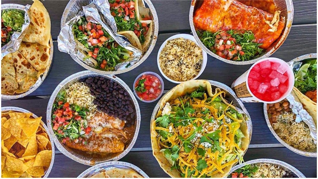 Overhead spread of tacos, burrito, salad, rice, chips and a drink in foil trays from Cafe Rio Fresh Modern Mexican