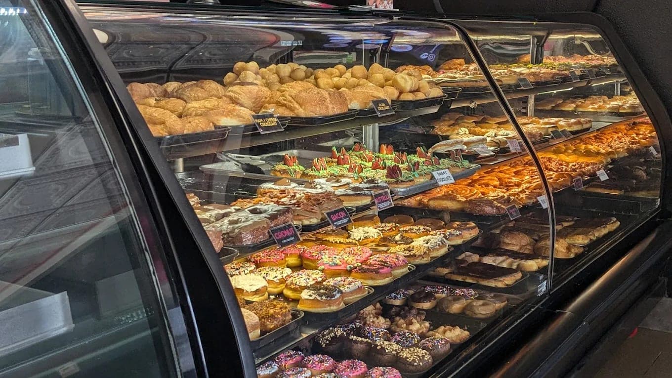 A curved bakery case filled with croissants on the upper shelf and rows of decorated donuts and pastries below