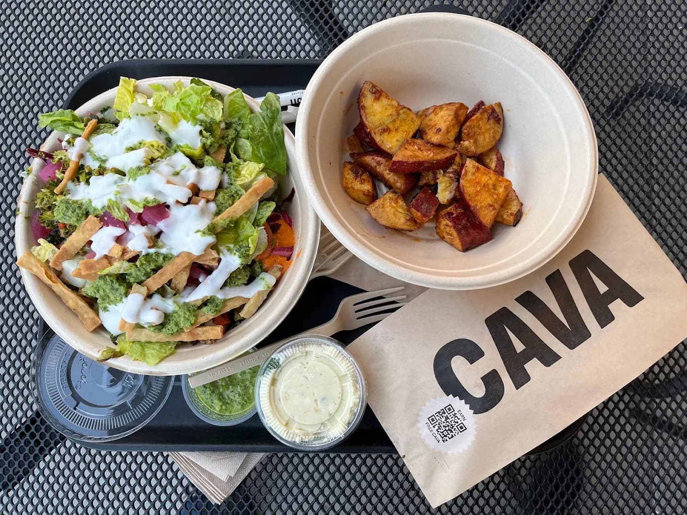 Salad bowl and roasted sweet potatoes on a tray beside a Cava paper bag on a patio table