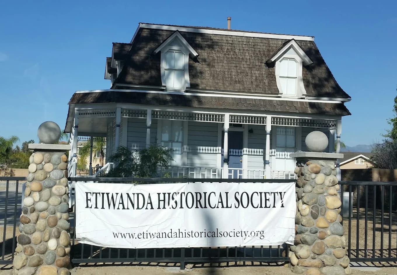 Two-story Victorian house with a front porch behind a stone-pillared fence and an Etiwanda Historical Society banner at Chaffey-Garcia House