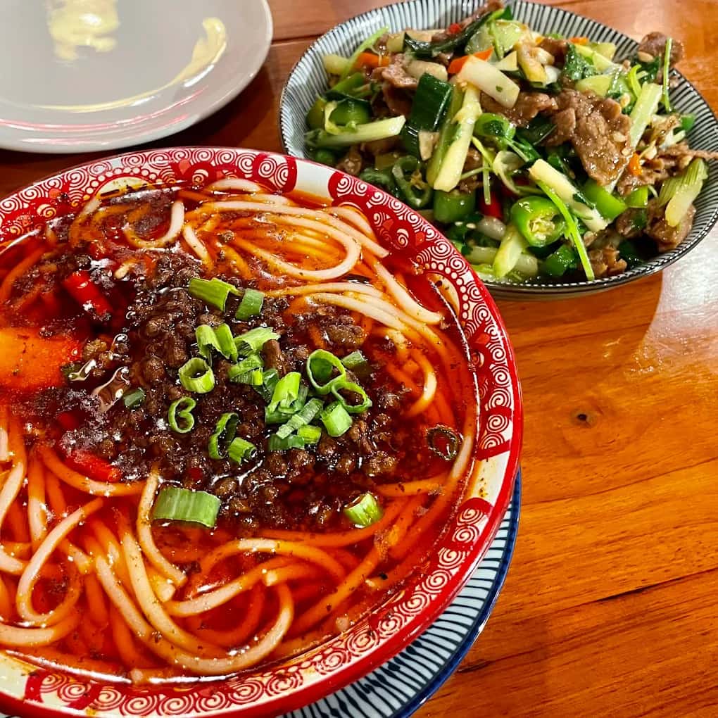Bowl of noodles in red chili broth topped with minced meat and scallions beside a beef and pepper stir-fry at Chili House