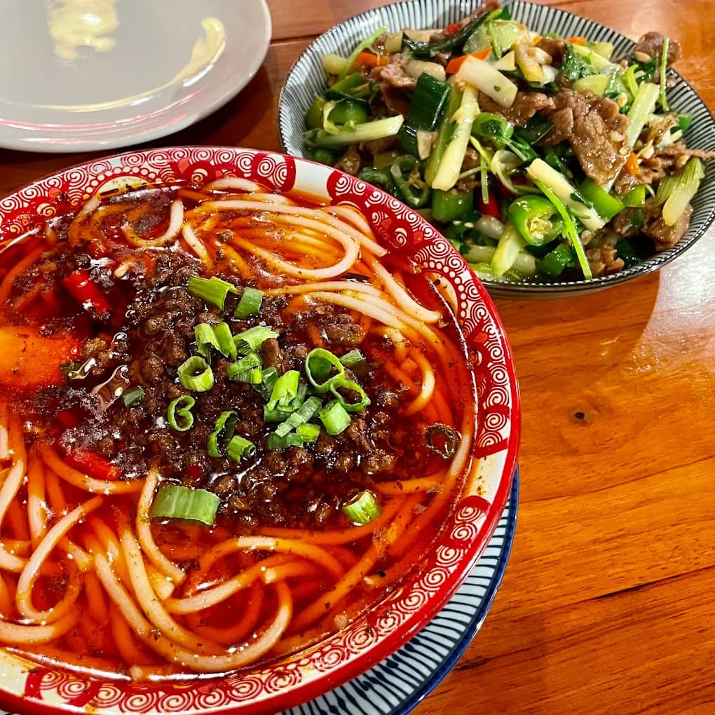 Bowl of noodles in red chili broth topped with minced meat and scallions beside a beef and pepper stir-fry at Chili House