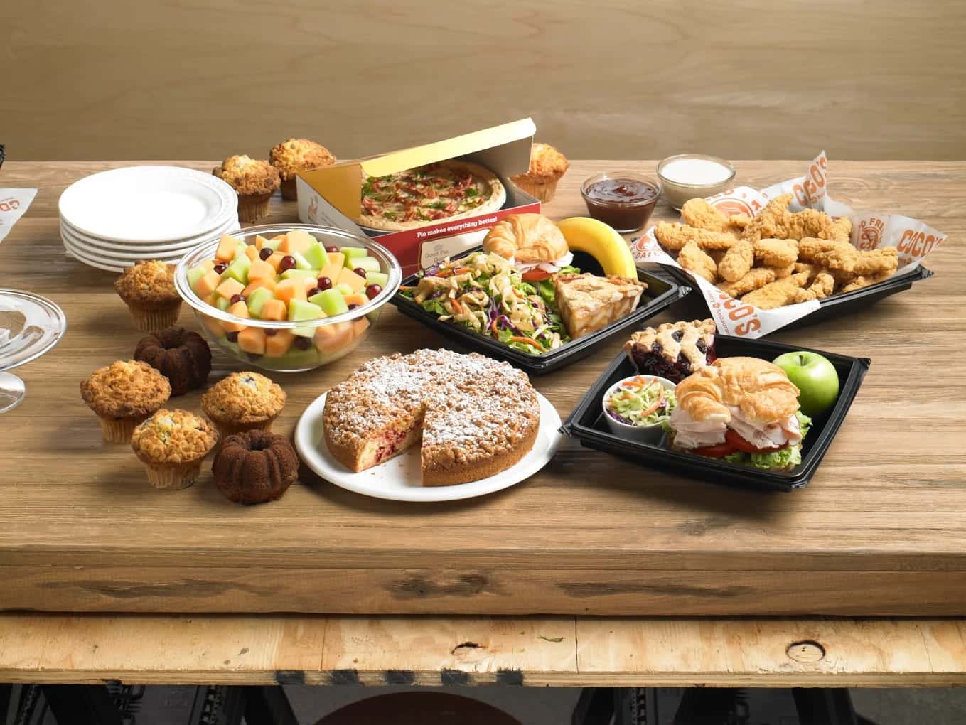 A spread of Coco's catering on a wooden table: muffins, a crumb-topped pie, fruit salad, salads, chicken tenders and croissant sandwiches