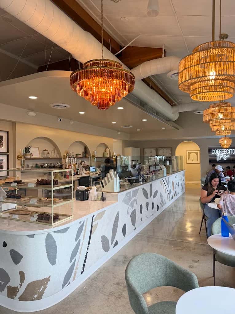Cafe interior at Coffeecito with a curved leaf-patterned counter, pastry case, rattan chandeliers and seated guests
