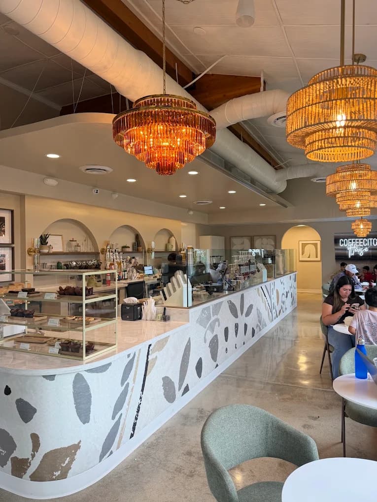 Cafe interior at Coffeecito with a curved leaf-patterned counter, pastry case, rattan chandeliers and seated guests