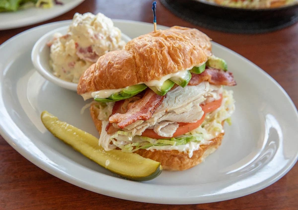A croissant sandwich with turkey, bacon, avocado and tomato, served with potato salad and a pickle spear