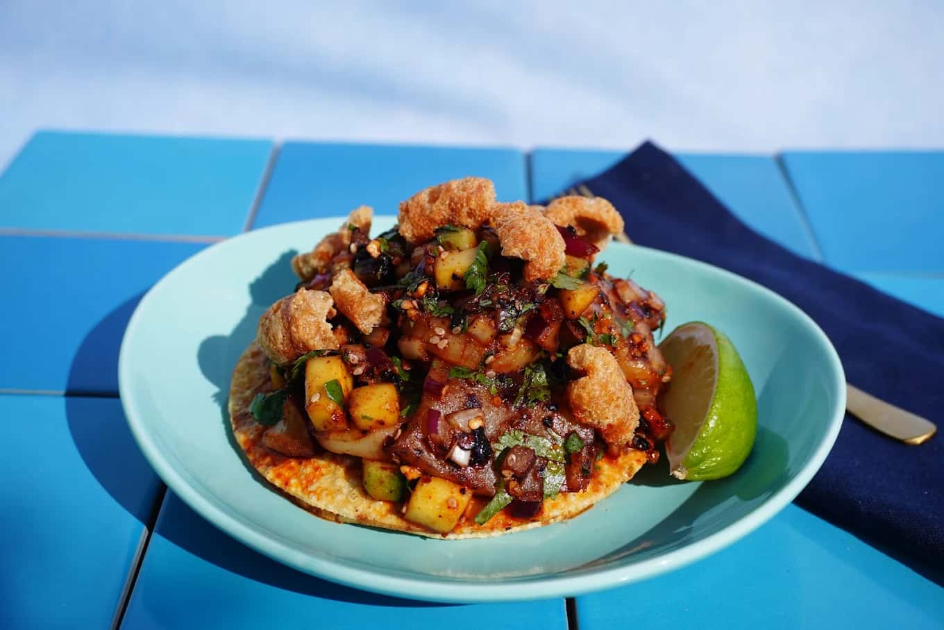 Tostada topped with diced seafood, mango, cilantro and chicharron with a lime wedge on a blue-tiled table at Costa de Oro