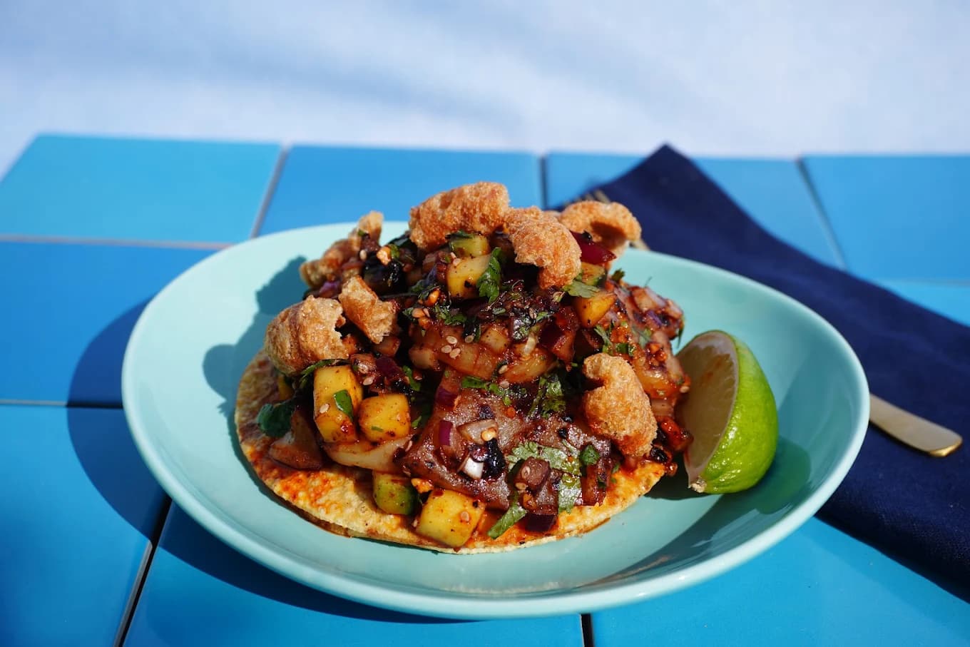 Tostada topped with diced seafood, mango, cilantro and chicharron with a lime wedge on a blue-tiled table at Costa de Oro