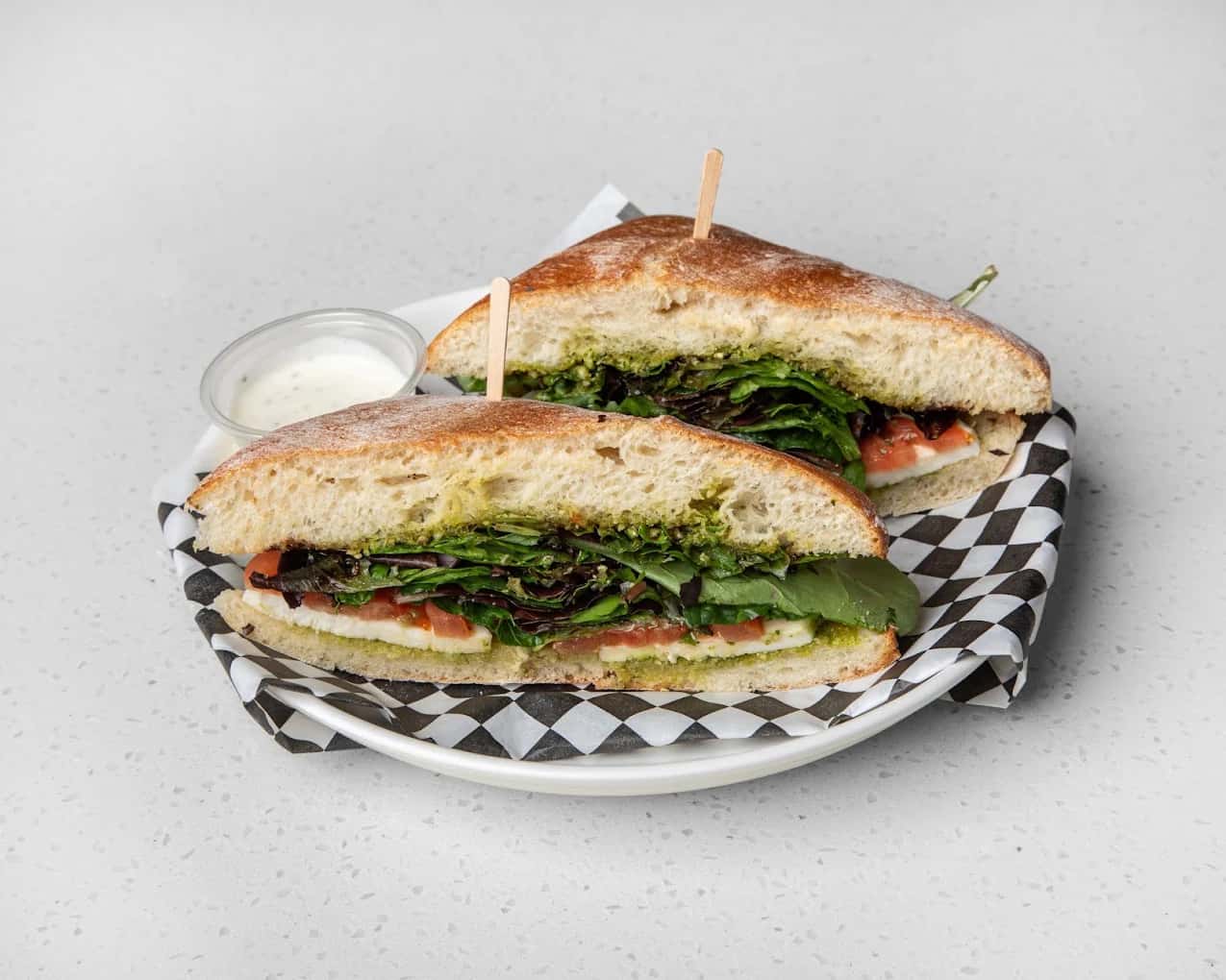Halved focaccia sandwich with mixed greens, tomato and mozzarella in a checkered-paper basket at Cotto & Crudo