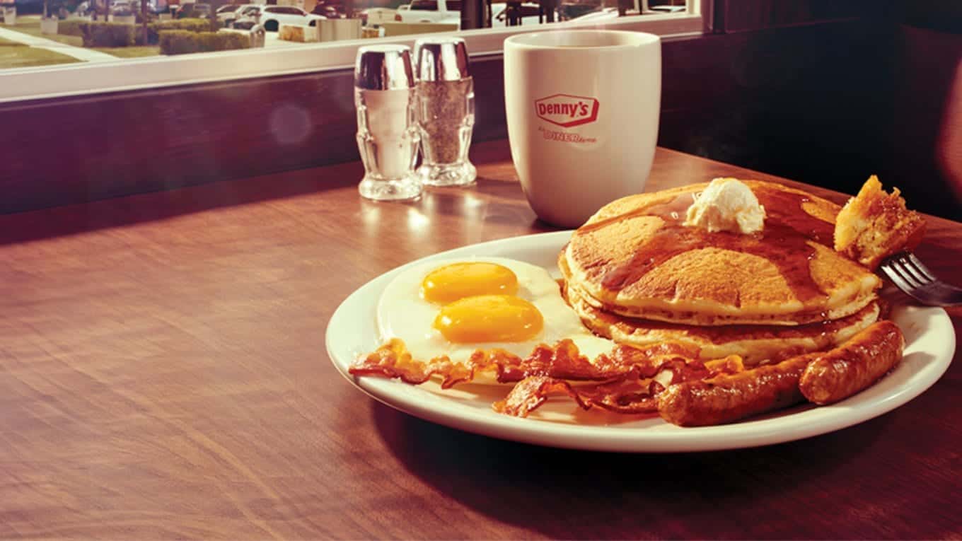A diner breakfast on a wooden table: pancakes with butter, fried eggs, bacon and sausage links beside a Denny's mug