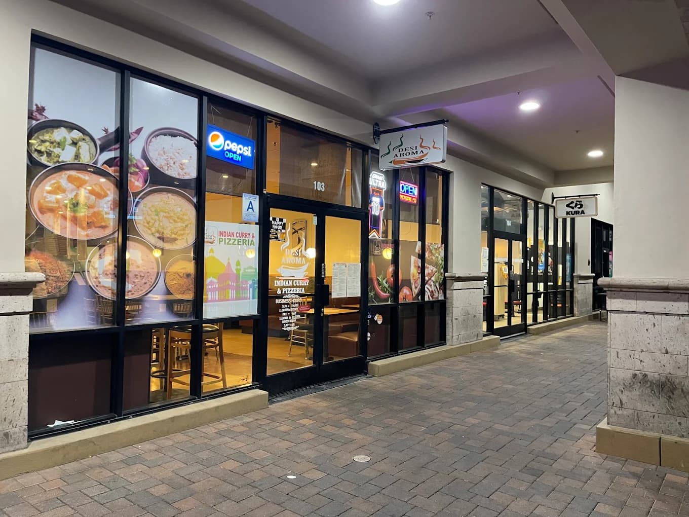 Storefront of Desi Aroma at night with Indian Curry & Pizzeria window graphics and a neon open sign