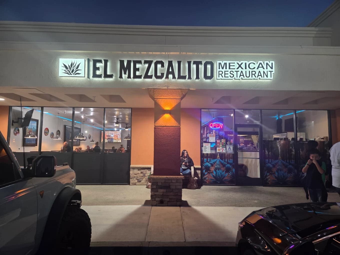 Lit storefront sign for El Mezcalito Mexican Restaurant at dusk with a neon open sign in the window