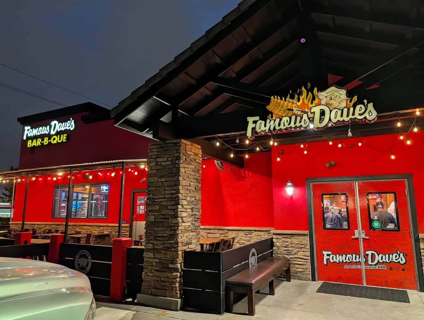The Famous Dave's entrance at night, red walls and stone pillars under string lights with benches beside the door