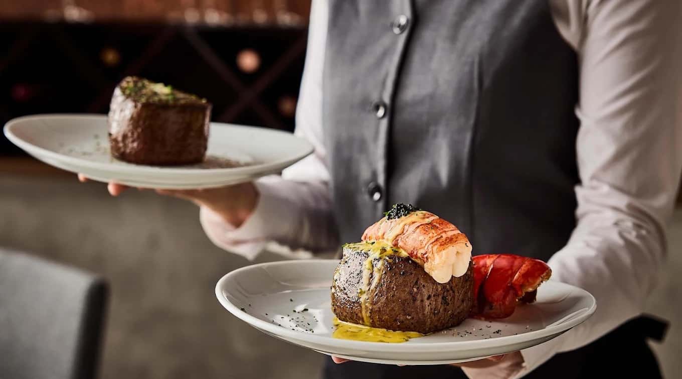 Server carrying plated filets, one topped with lobster tail, at Fleming's Prime Steakhouse & Wine Bar