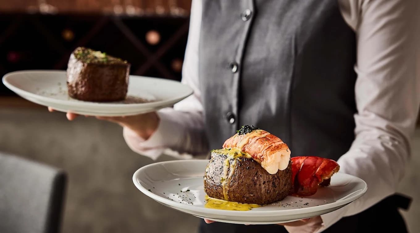Server carrying plated filets, one topped with lobster tail, at Fleming's Prime Steakhouse & Wine Bar