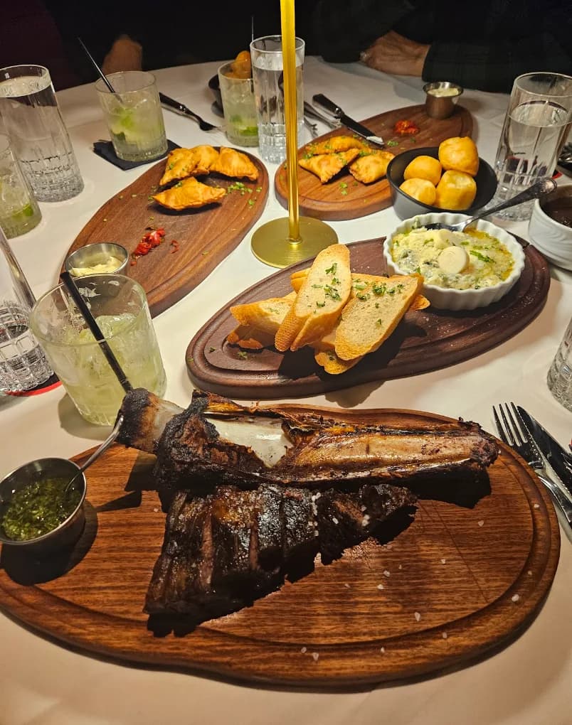 Table spread with a bone-in steak, empanadas, bread and cheese rolls at Fogo de Chão