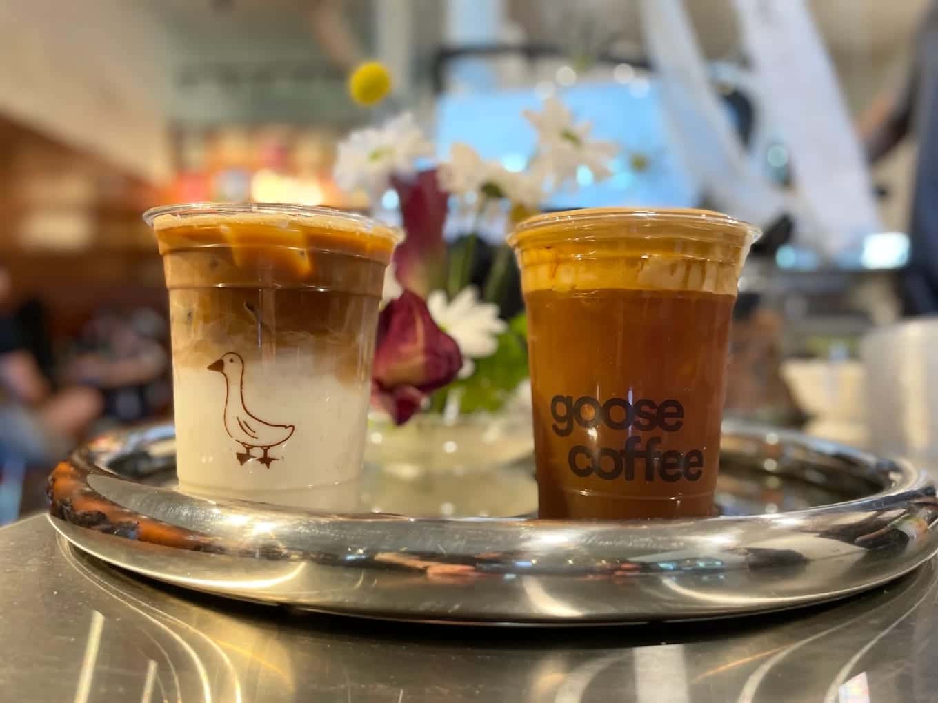 Two iced coffees on a steel tray at Goose Coffee, one layered cup showing the goose logo and one branded cup beside a vase of flowers