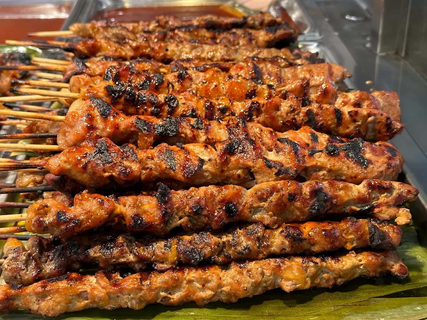 Grilled pork skewers stacked on a banana leaf in a warming tray, charred along their edges