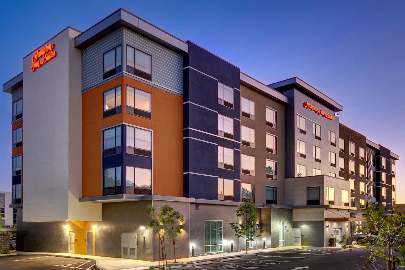 Exterior of the Hampton Inn & Suites Ontario Rancho Cucamonga at dusk, with orange, gray and navy panels and lit signs