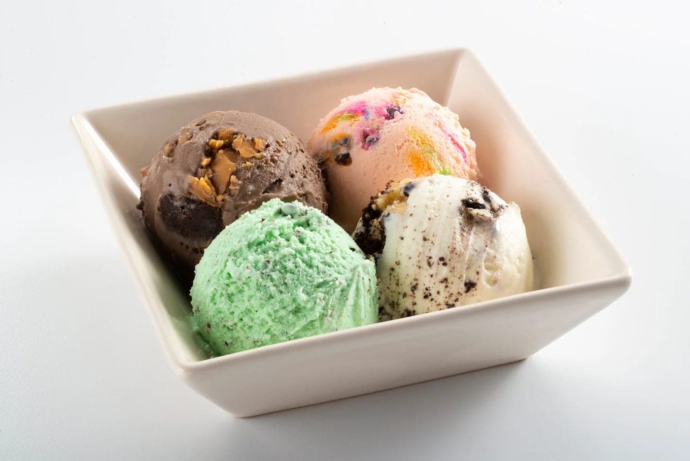Four scoops in a square white bowl: chocolate with nuts, a rainbow flavour, mint and cookies and cream