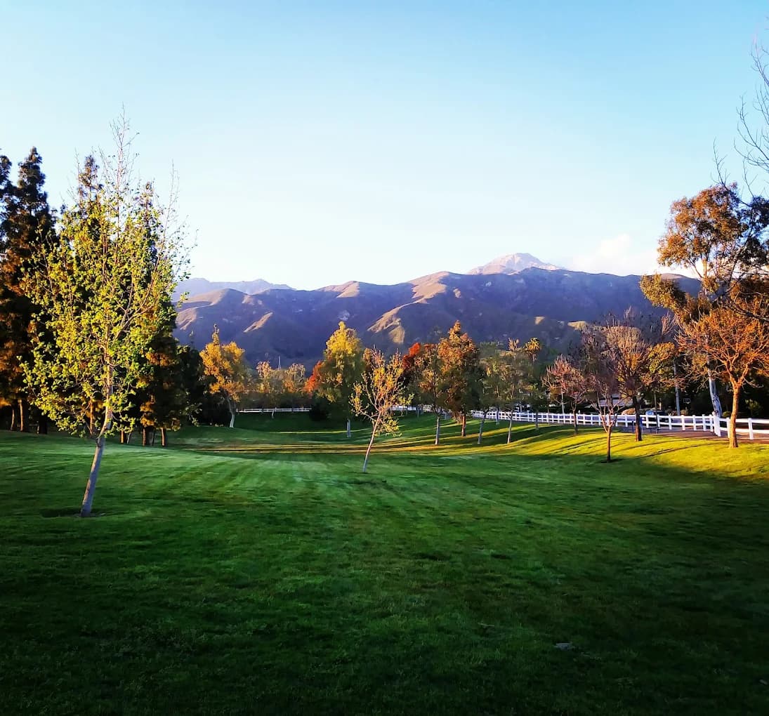 Grass lawn, trees and a white rail fence at Heritage Community Park with mountains behind