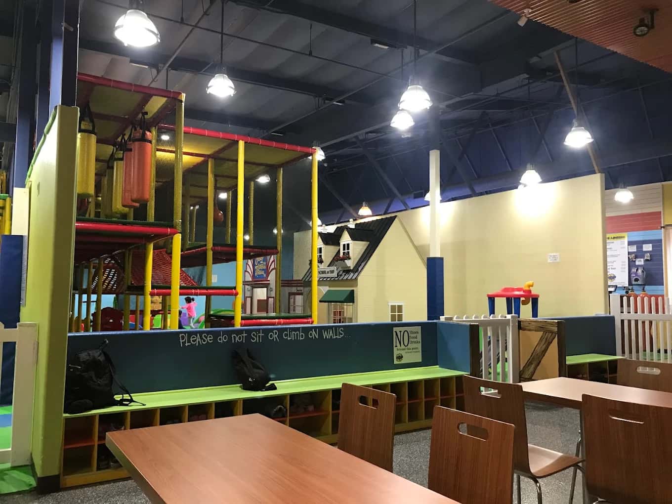 Indoor play structure with padded posts and a playhouse beside tables and shoe cubbies at House of Play
