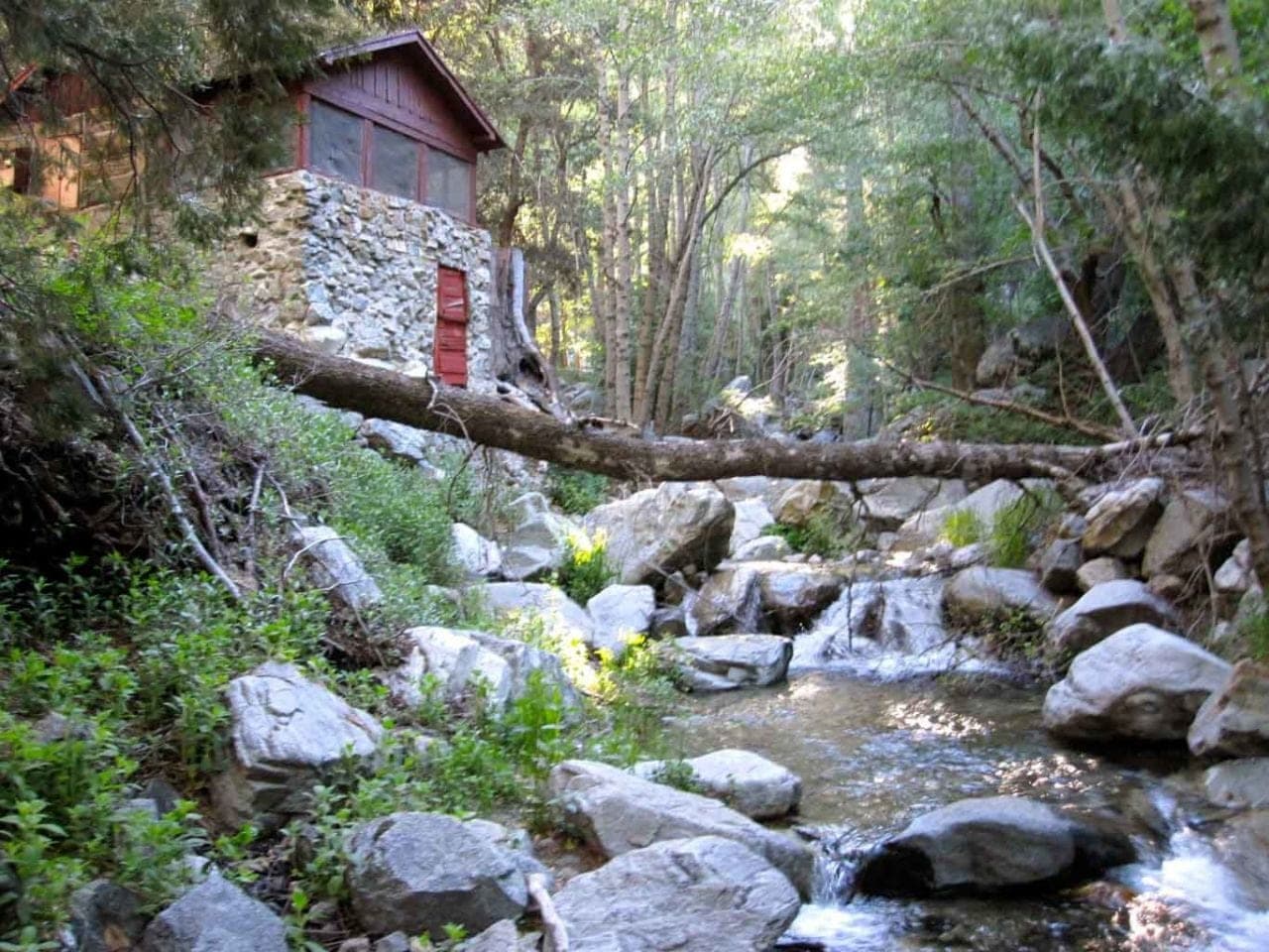 A stone cabin with a red door above the creek in Icehouse Canyon, a fallen log spanning the boulders below it