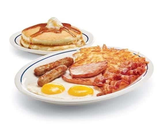 A breakfast platter of fried eggs, sausage links, ham, bacon and hash browns with a short stack of pancakes behind