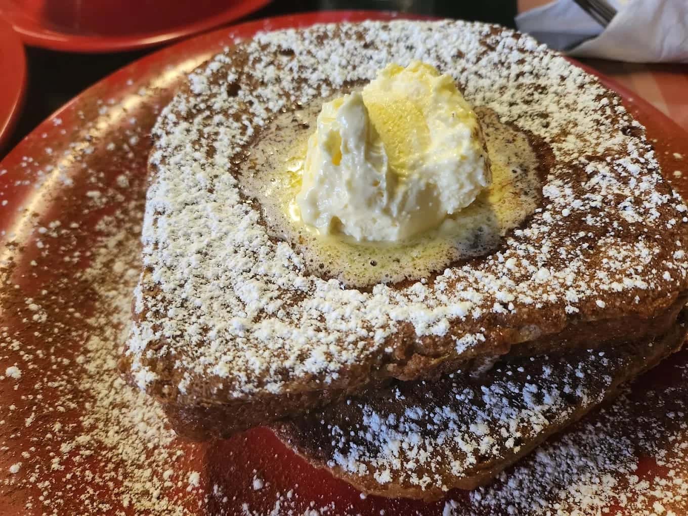 Thick French toast under a heavy dusting of powdered sugar, topped with whipped cream on a red plate