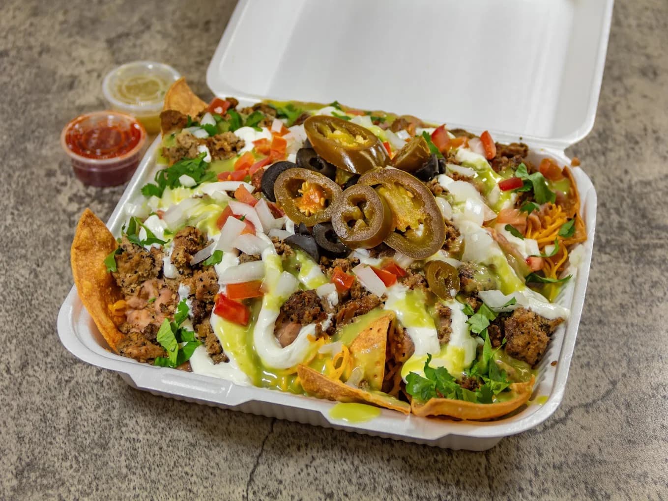 Takeout box of loaded nachos from Juanita's III Rancho Cucamonga, topped with jalapeños, olives, sour cream and green salsa