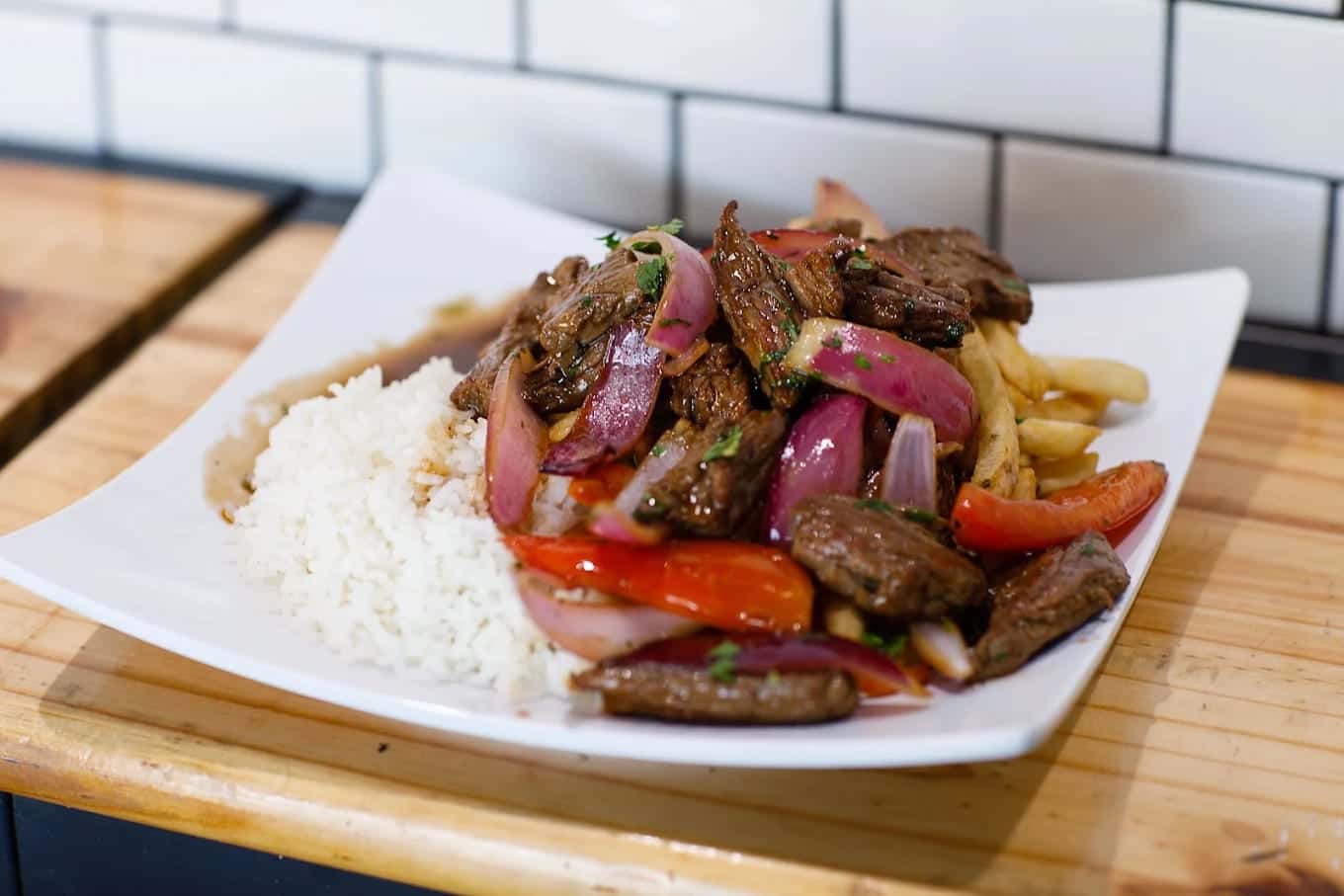 Lomo saltado on a square plate: sautéed beef with red onion, tomato and fries alongside white rice