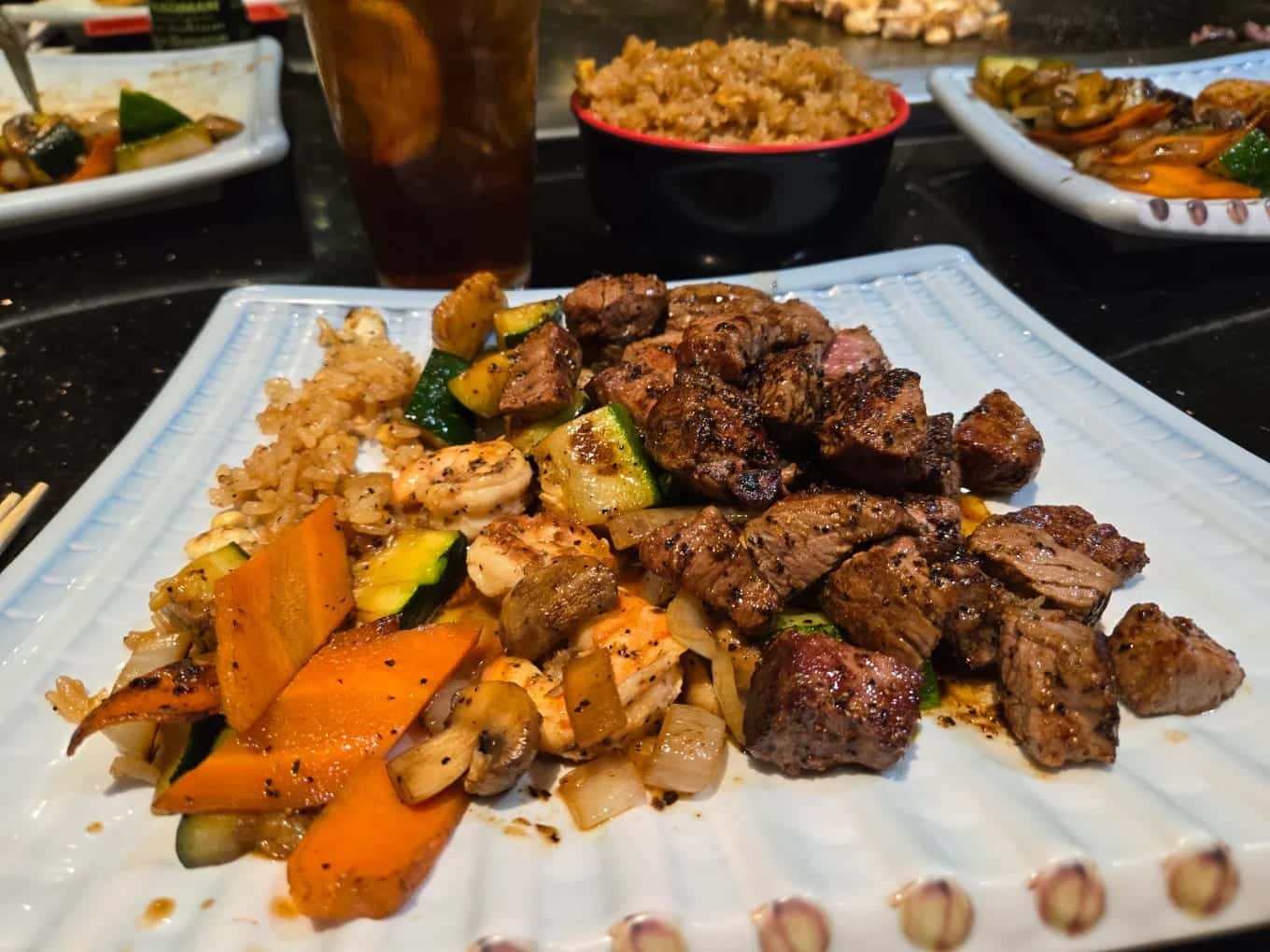 A hibachi plate of steak, zucchini, carrots, mushrooms and onion with fried rice, an iced tea behind it